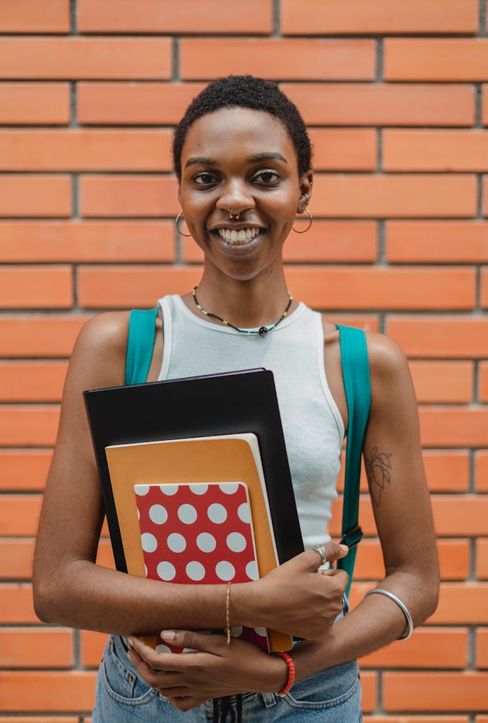 Cheerful young black female with short hair in casual outfit standing against brick wall with folder and copybooks looking at camera